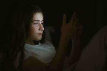 child teenager girl lies in bed in evening or at night and looks into phone. mobile addiction.