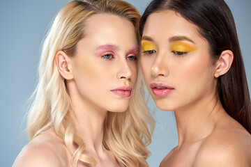 portrait of two interracial women with bold makeup posing on blue background, vibrant eye shadows