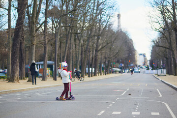Adorable preschooler girl riding her scooter in a city park on sunny spring day