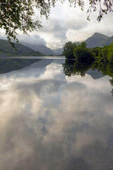 Bright sunny clouds, mountains and forest reflected on the calm tranquil surface of a vast lake. Llyn Padarn, Llan Beris, Snowdonia Wales.