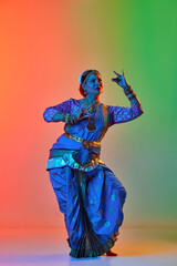 Fototapeta premium Odissi dance. Beautiful mature indian woman in traditional dress dancing indian dance against gradient studio background in neon light. Concept of beauty, fashion, India, traditions, choreography, art