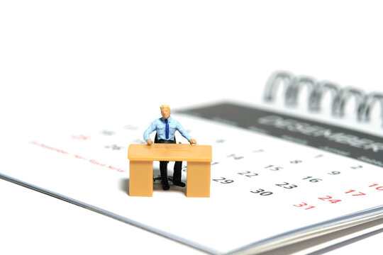 Miniature tiny people toy figure photography. Due date concept. A businessmen seat on his desk, above calendar. Isolated on a white background