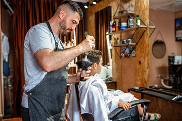 Barber puts talcum powder on the brush. Little boy patiently sits on the char in barber shop in the background.  