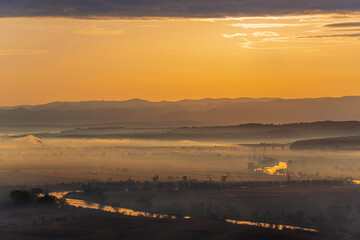Aerial view of the valley in early morning mist, beautiful in the highlands. Low clouds and fog cover the sleeping meadow. Alpine mountain valley mists landscape at dawn. Serene moment in rural area