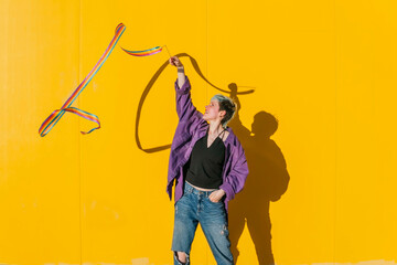 Lesbian woman waving rainbow colored ribbon in front of yellow wall