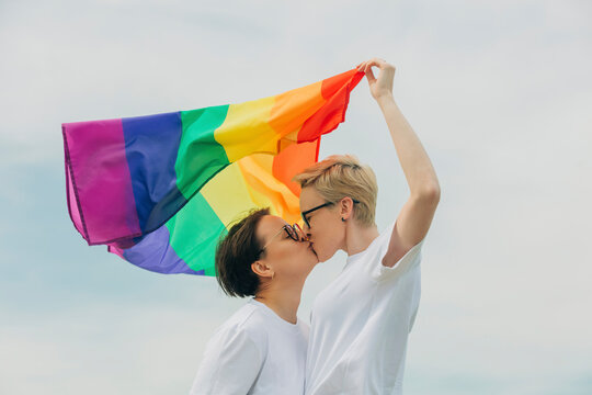 Loving lesbians with rainbow flag kissing each other under sky