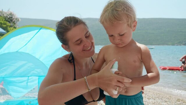 Loving young mother diligently applying sunscreen spray to her baby son on a sunny beach. Concept of healthcare and family holidays.