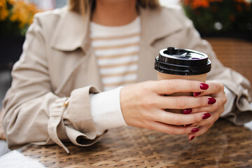 Close-up of a woman holding a cup of coffee in a street cafe, in a beige trench coat and a cozy...