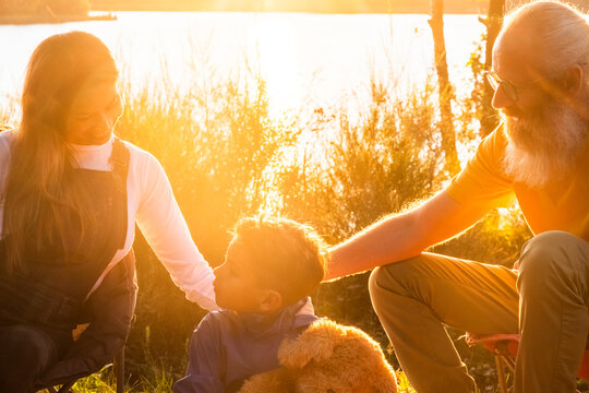 As the golden sun sets over the tranquil forest lake, a beautiful scene unfolds. A young Asian mother and her older bearded Caucasian husband, together with their mixed-race son, have embarked on a