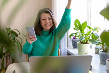 Happy young woman rejoicing over victoring on internet, completing project, done or new job, success, winning online lottery, finishing mortgage. Female glad at computer in home office with  phone