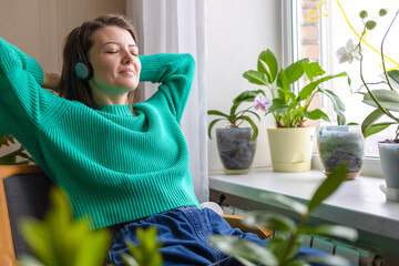 young woman takes break from hustle and bustle, listening music, audiobook on wireless headphones in cozy green house with plants