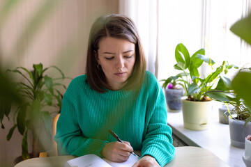 Serious young woman working, planning in workspace in green cozy home in plants. female writing strategy, business plan, ideas in home office. planning concept