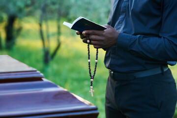 Close-up of African American man in priest attire holding rosary beads and open Holy Bible while standing in front of coffin during burial service