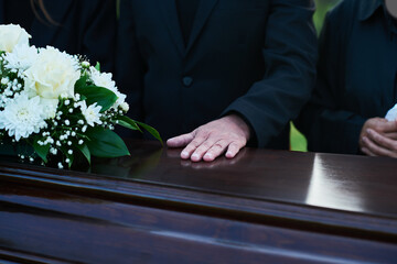 Close-up of mature man keeping hand on top of closed lid of coffin with dead body of relative or...