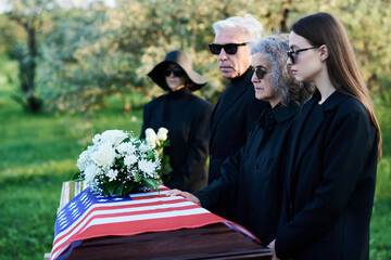 Row of family members in sunglasses and mourning clothes standing by coffin covered with American flag and lamenting their dead relative