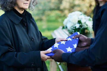 Mature grieving widow in black attire taking folded stars-and-stripes flag from hands of African...