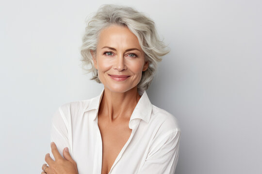 Attractive sixty-year-old female artist with gray hair and a warm look, wearing a widely unbuttoned white shirt, posing against a neutral background.