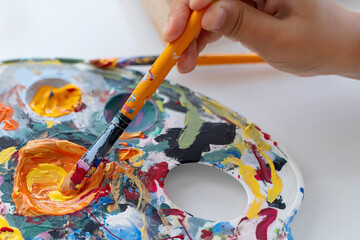 Artist's hand picks up acrylic or oil paint from palette on brush. Closeup paintbrush picking orange color from an artist palette. Colorful image from an artist’s studio or a school showing creative e