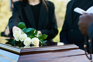 Young woman in black suit putting four fresh white roses on closed lid of coffin while standing by...