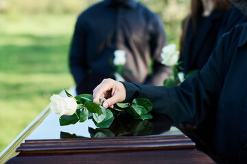 Hand of mature woman in mourning attire putting white rose on top of closed coffin lid while standing in front of camera against other people