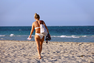 Tanned woman in white bikini walking towards the sea on sandy beach. Vacation at ocean coast