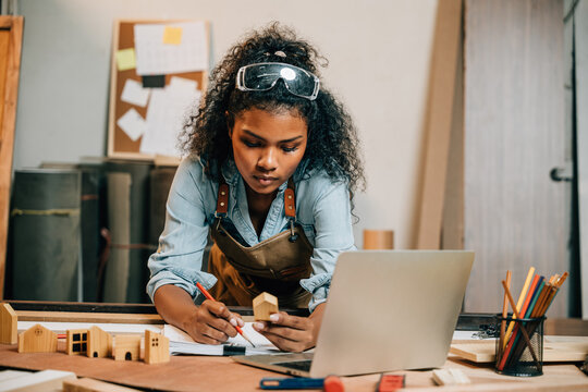 Carpenter America Black Woman Curly Hair Sketch Making Notes In Work Paper While Standing At Desk With Laptop Computer In Wood Workshop, Young Female Working Learning Online At Woodshop