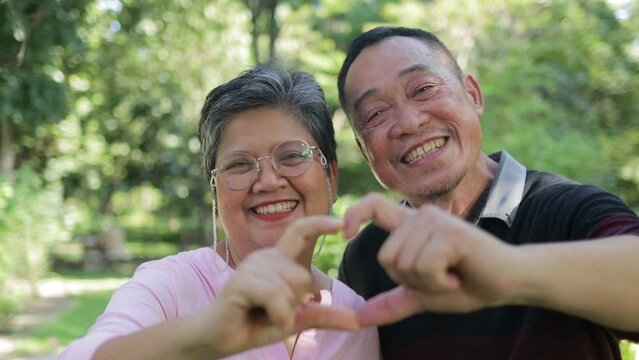 Portrait of an elderly Asian couple smiling happily in the park. They both made their hands into a heart shape. Living happily in retirement