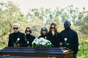 Group of intercultural grieving people in mourning attire and sunglasses holding white roses while...