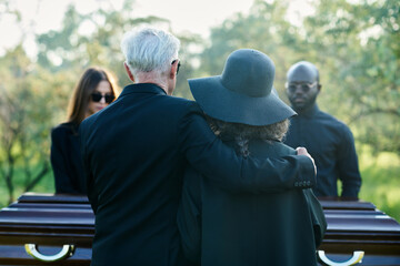 Rear view of mature grey haired man embracing his wife in black hat while both standing in front of...