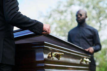 Two intercultural men in black attire closing coffin lid after farewell ceremony before carrying it...