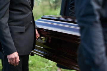 Focus on man in black suit holding by handle of wooden coffin while carrying it together with other...