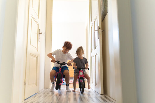 Siblings are riding bicycles indoors, in the hallway of the house.