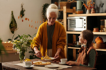 Senior woman putting piece of homemad apple pie on plate of her cute granddaughter sitting by table...