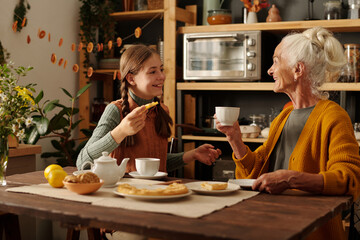 Cheerful girl with piece of apple pie looking at her granny with cup of tea while both sitting by...