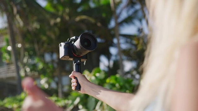 Female travel blogger films video on ocean beach. Blonde woman shoots blog walking with pro camera on tropical island. Influencer records story for social network. Vlogger works on movie, back view.