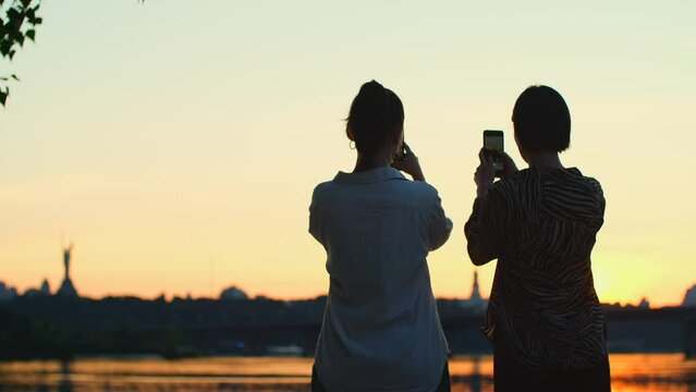 Two women take photos of river with mobile phone. Beautiful girls taking video of sunset on smartphone, female friends relaxing on the beach
