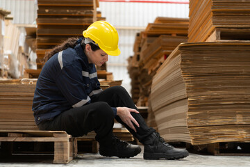 Warehouse worker sitting on the floor in front of stack of cardboard boxes with pain in his leg from hitting wooden pallets.