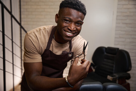 African American barber in working apron is leaning on barber chair