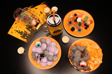 Black table ready to start a terrifying Halloween party celebration. 3 orange plates full of colorful sweets and candies. Jug full of orange soda. 5 lit candles. 3 napkins and a jar with confectionery