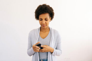 Studio portrait of attractive curly African American woman using smartphone typing browsing looking to device screen. Focused black female holding mobile cellphone on isolated white background.