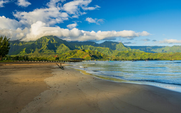 Panorama of the sandy beach of Hanalei Bay with pier and Na Pali mountains shrouded in sunlit clouds