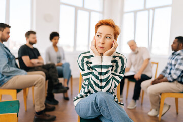 Portrait of pensive young woman covering ears with hands and looking away sitting in circle at group psychological counseling. Concept of mental health, psychotherapy, social issues, support.