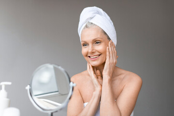 Beautiful mature woman with towel on head looking at mirror, using new brand anti-aging cosmetics, home interior