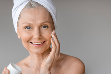 Daily treatments. Beautiful caucasian mature woman holding jar of cream and applying moisturizer on cheek, free space
