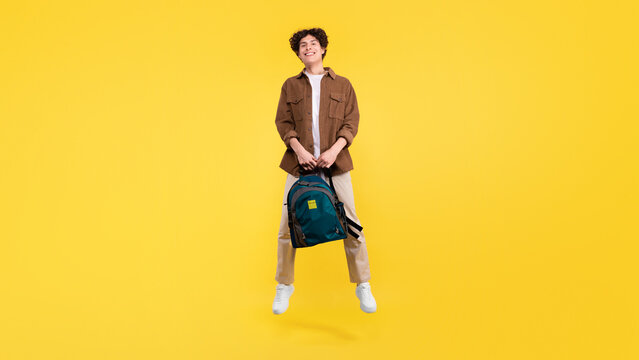 Happy student guy with backpack jumping over yellow studio background