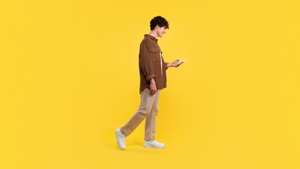 Young man using smartphone texting while walking on yellow background