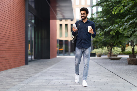 Cheerful arab guy going to office, using smartphone, drinking coffee