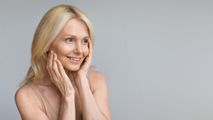 Beautiful mature woman looking aside at copy space and touching cheeks, enjoying skincare treatments, panorama