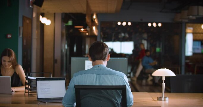 A guy working at his desk in a modern office space, facing a large monitor, with a colleague sitting across from him