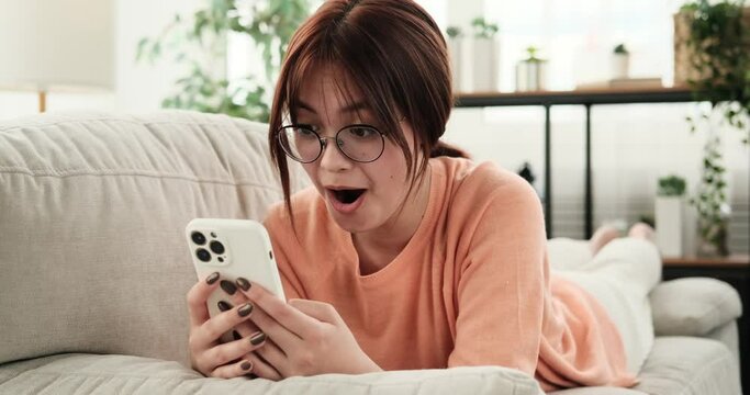 Lying on the couch, a teenage girl using her phone suddenly becomes greatly surprised, her eyes widening in astonishment. The unexpected information on the phone screen triggers a strong reaction.